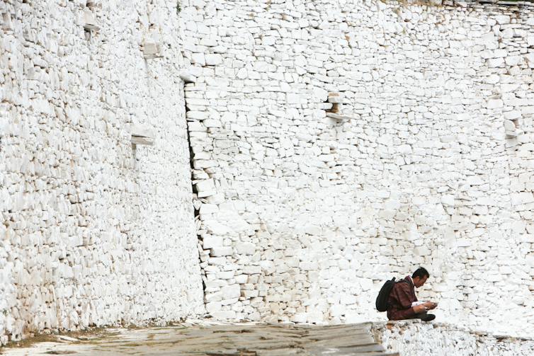 Bhutanese man checking his mobile phone next a white stone wall.